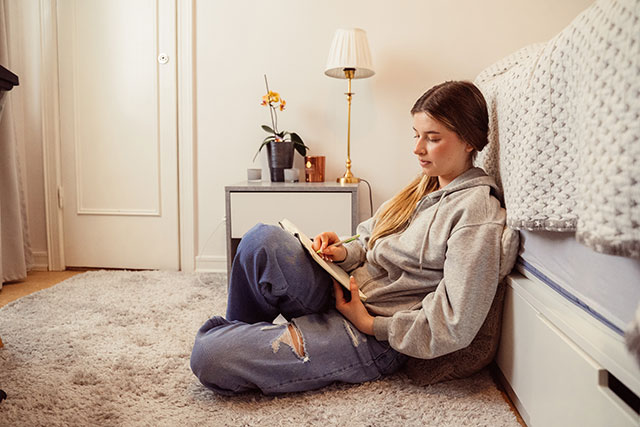 Maria sitter på golvet. lutande mot sängen och skriver i en bok